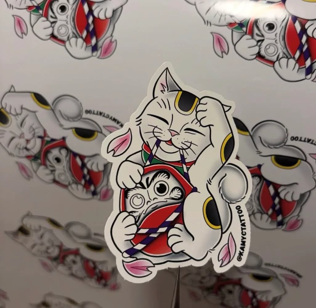 Lucky cat sticker by Kamy Cao — a smiling maneki-neko hugging a red daruma, with cherry blossom petals, in Japanese tattoo style.