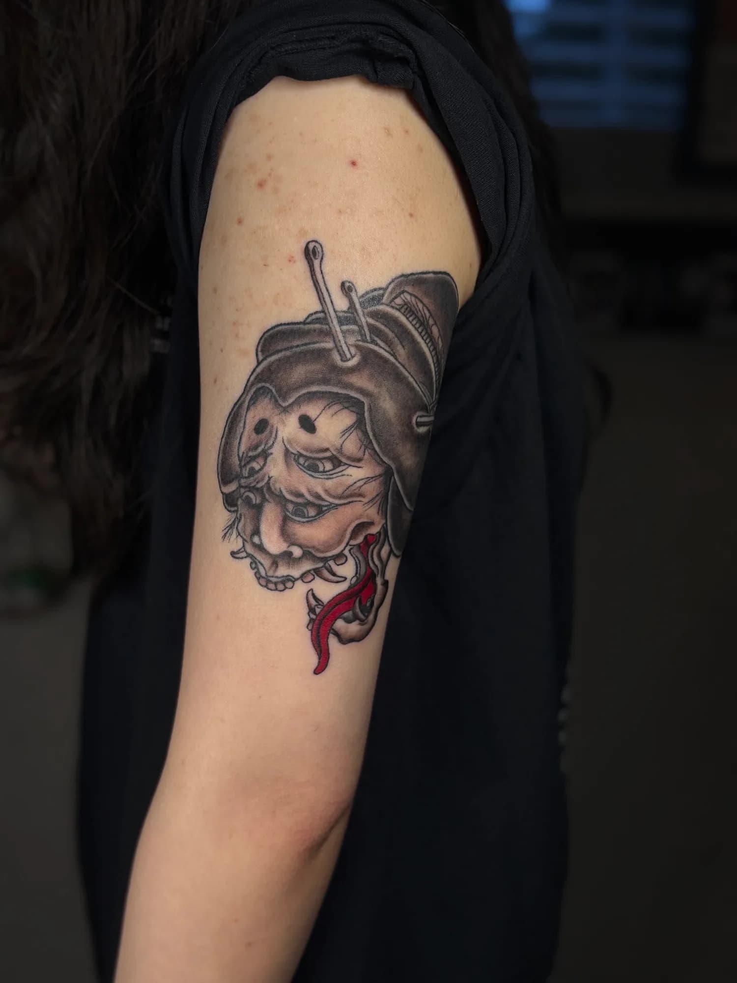 Black and grey Japanese yokai mask tattoo with a red tongue, on the shoulder, by Kamy Cao.
